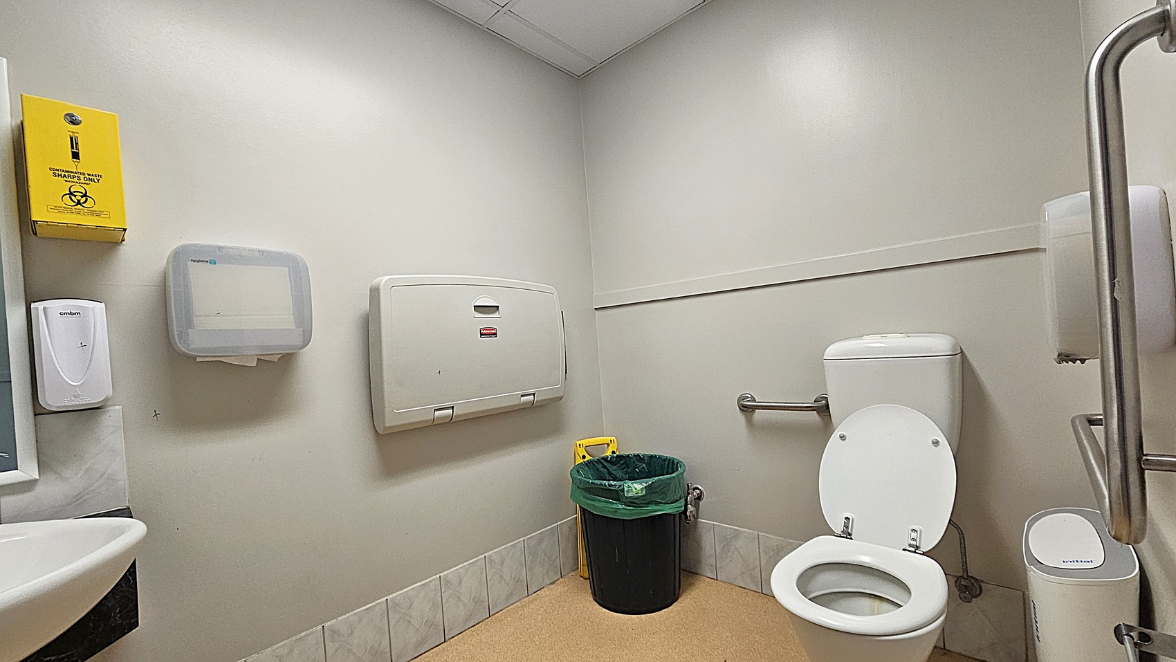 A public restroom with a white toilet and open lid on the right, equipped with a grab bar. A black bin with a green liner is next to the toilet. On the left wall, there is a yellow sharps disposal box, a soap dispenser, a paper towel dispenser, and a baby changing station. The floor is covered with beige tiles, and the walls are light-coloured. The ceiling is made of white tiles.