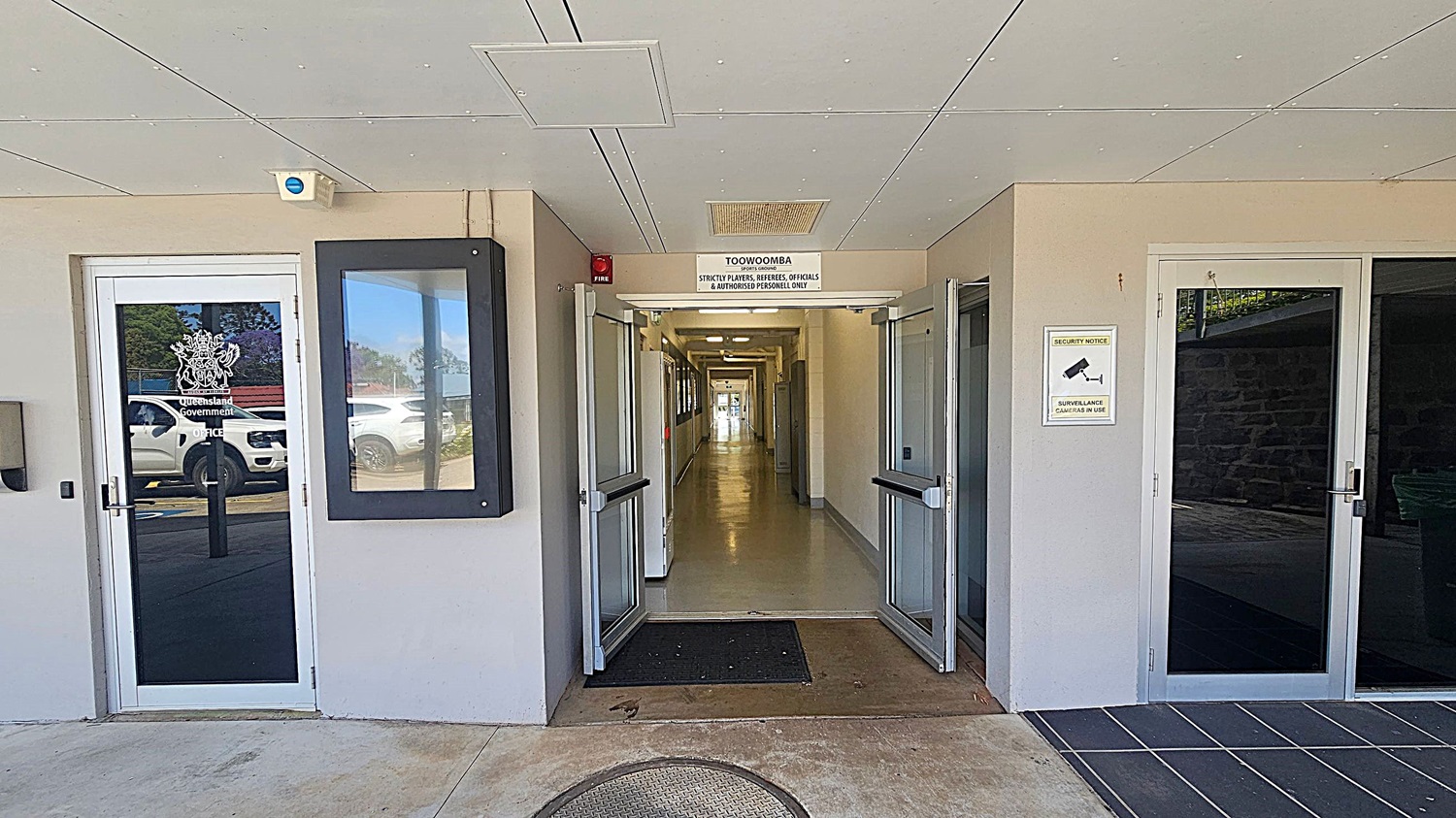 A corridor entrance with glass doors on both sides. The left door has a Queensland Government logo and reflects parked cars. The right door has a security notice about surveillance cameras. In the centre, double doors lead to a long hallway with a sign above reading "TOOWOOMBA STRICTLY PLAYERS, REFEREES, OFFICIALS & AUTHORISED PERSONNEL ONLY". The ceiling is white with panels, and the floor is tiled.