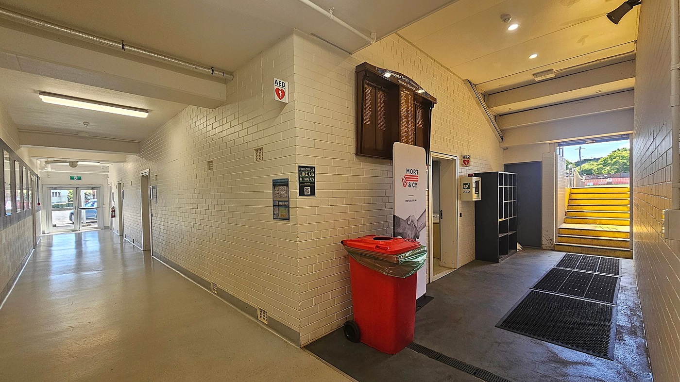 A corridor with white brick walls and a light-coloured floor. On the left, there are windows and a glass door leading outside, with a car visible through the door. On the right, a red bin and a sign are placed against the wall. Above them, a wooden honours board is mounted. Further right, a set of yellow stairs leads upwards, with a small black shelving unit nearby. The ceiling has fluorescent lights and exposed pipes.