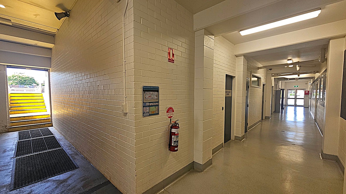 A corridor with white brick walls and a polished floor. On the left, there's an open doorway leading outside with yellow steps visible. A fire extinguisher is mounted on the wall near a sign. On the right, several doors line the hallway, including one labelled "Conference Room." The ceiling has fluorescent lights, and the corridor extends towards a glass door at the far end, allowing natural light to enter.