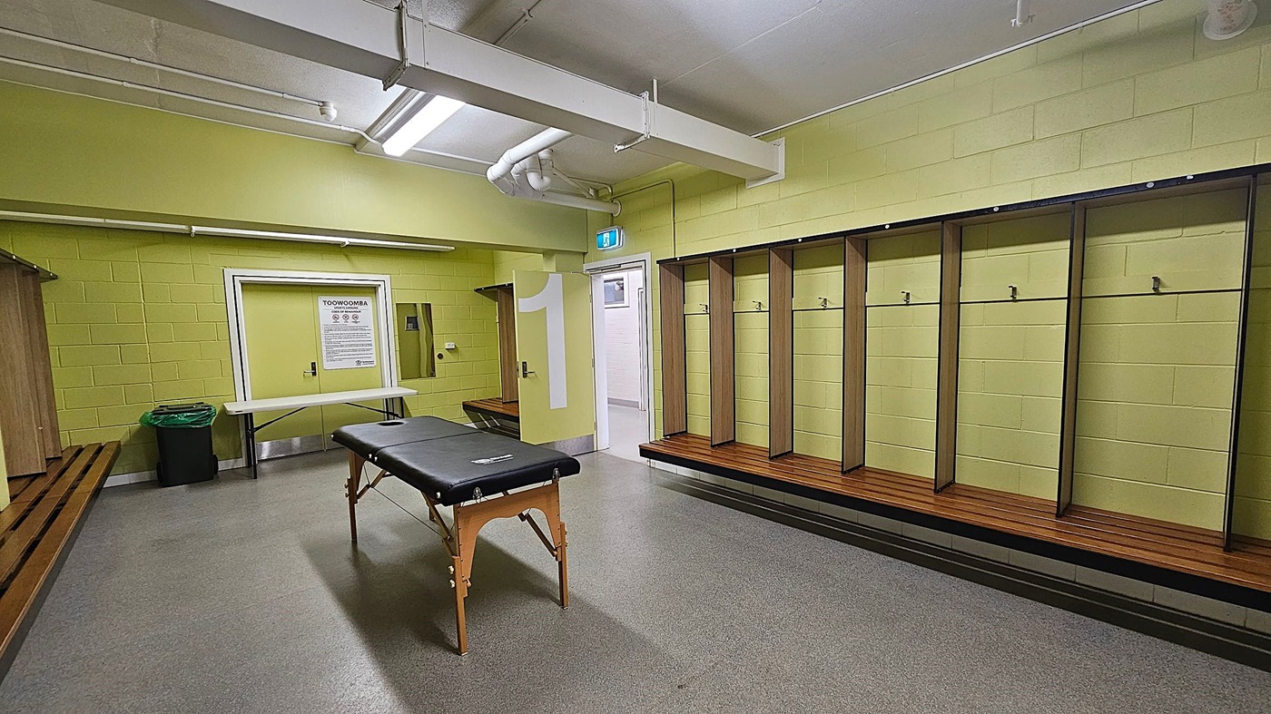 A locker room with yellow brick walls and a grey floor. On the right, there are wooden benches with open lockers above them, each with a hook. In the centre, a black massage table stands on wooden legs. On the left, a long wooden bench runs along the wall, with a green bin nearby. A white door with a large number "1" and a sign above it is visible in the background. The ceiling has exposed pipes and fluorescent lights.