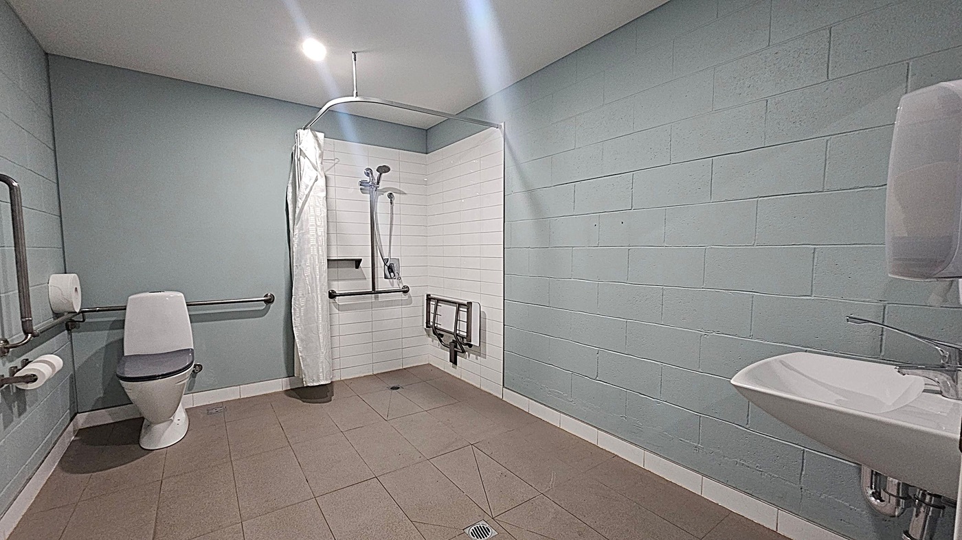A spacious, accessible bathroom with a beige tiled floor and white brick walls. On the left, there's a toilet with grab bars and a toilet roll holder. In the centre, a shower area with a curtain, handheld showerhead, and a foldable seat. On the right, a white sink with a lever tap and a paper towel dispenser. The ceiling is plain with a recessed light.