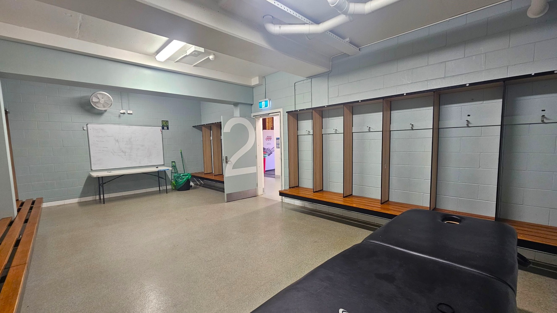 A locker room with grey walls and a beige floor. On the right, there are wooden benches with open lockers above them. In the centre, a door marked with a large number "2" is open, revealing a glimpse of another room. On the left, a whiteboard is mounted above a folding table, with a fan on the wall above. The ceiling has exposed pipes and fluorescent lights. A green bag and cleaning equipment are near the table.