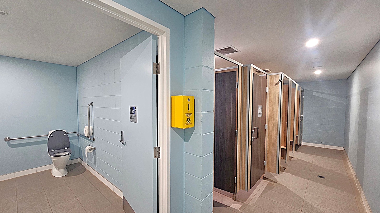 A public restroom with light blue walls and beige tiled flooring. On the left, there's an accessible toilet with grab bars and a toilet paper holder. A yellow sharps disposal box is mounted on the wall near the entrance. On the right, there are several wooden cubicle doors, each with a handle and a sign. The ceiling is white with recessed lighting.