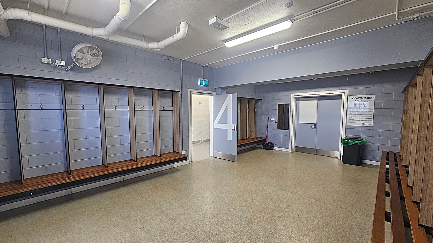A locker room with light grey walls and a beige floor. On the left, there are wooden benches with hooks above them. In the centre, a door marked with a large number "4" is open, leading to another room. On the right, more benches and a closed door with a sign above it. A green bin is near the door. The ceiling has exposed pipes and fluorescent lights.