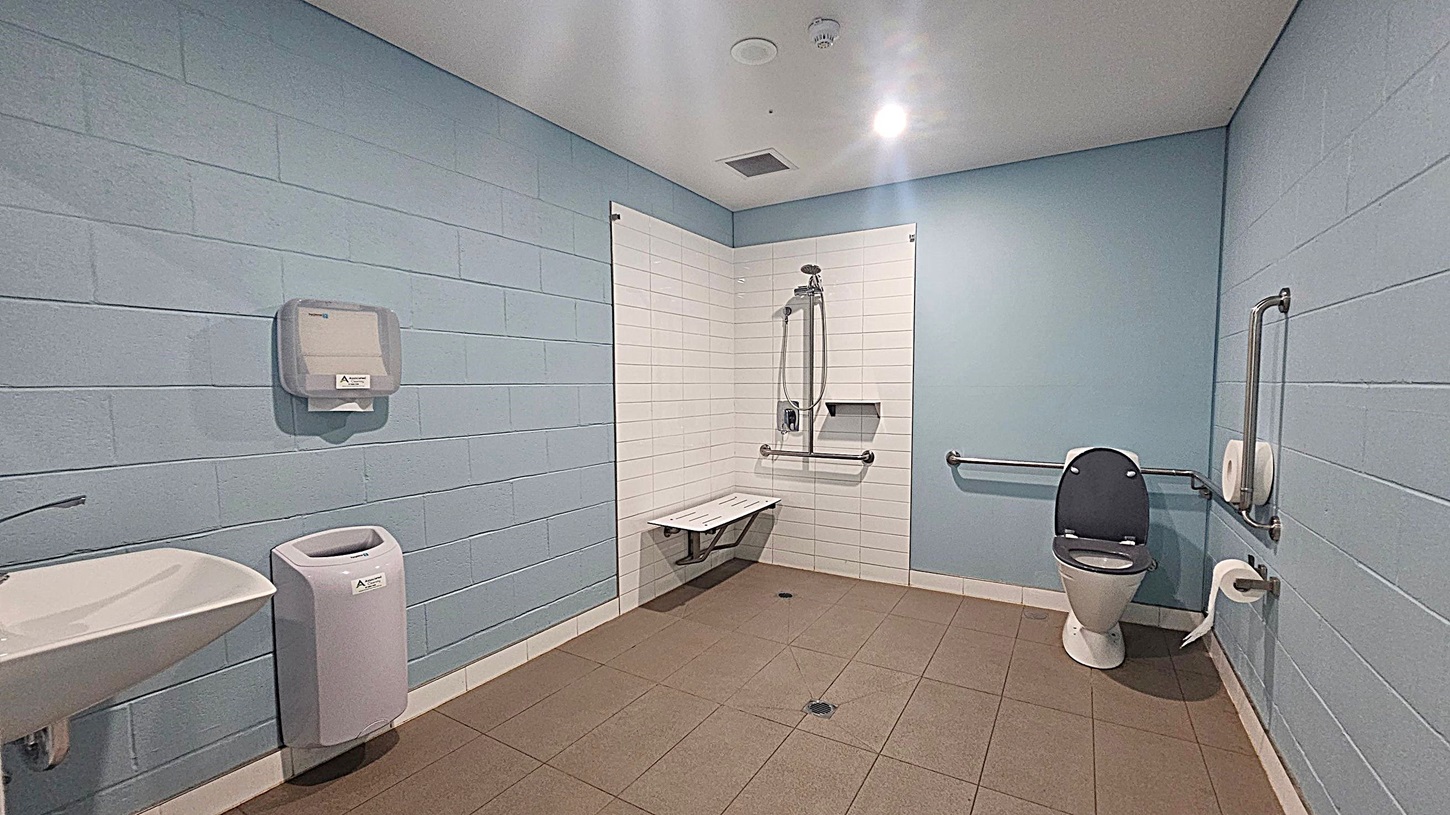 A spacious accessible bathroom with light blue walls and a grey tiled floor. On the left, there's a white sink with a hand dryer above it. In the centre, a shower area with white tiles features a foldable seat and grab bars. On the right, a toilet with a black seat is equipped with grab bars and a toilet roll holder. The ceiling is white with recessed lighting.