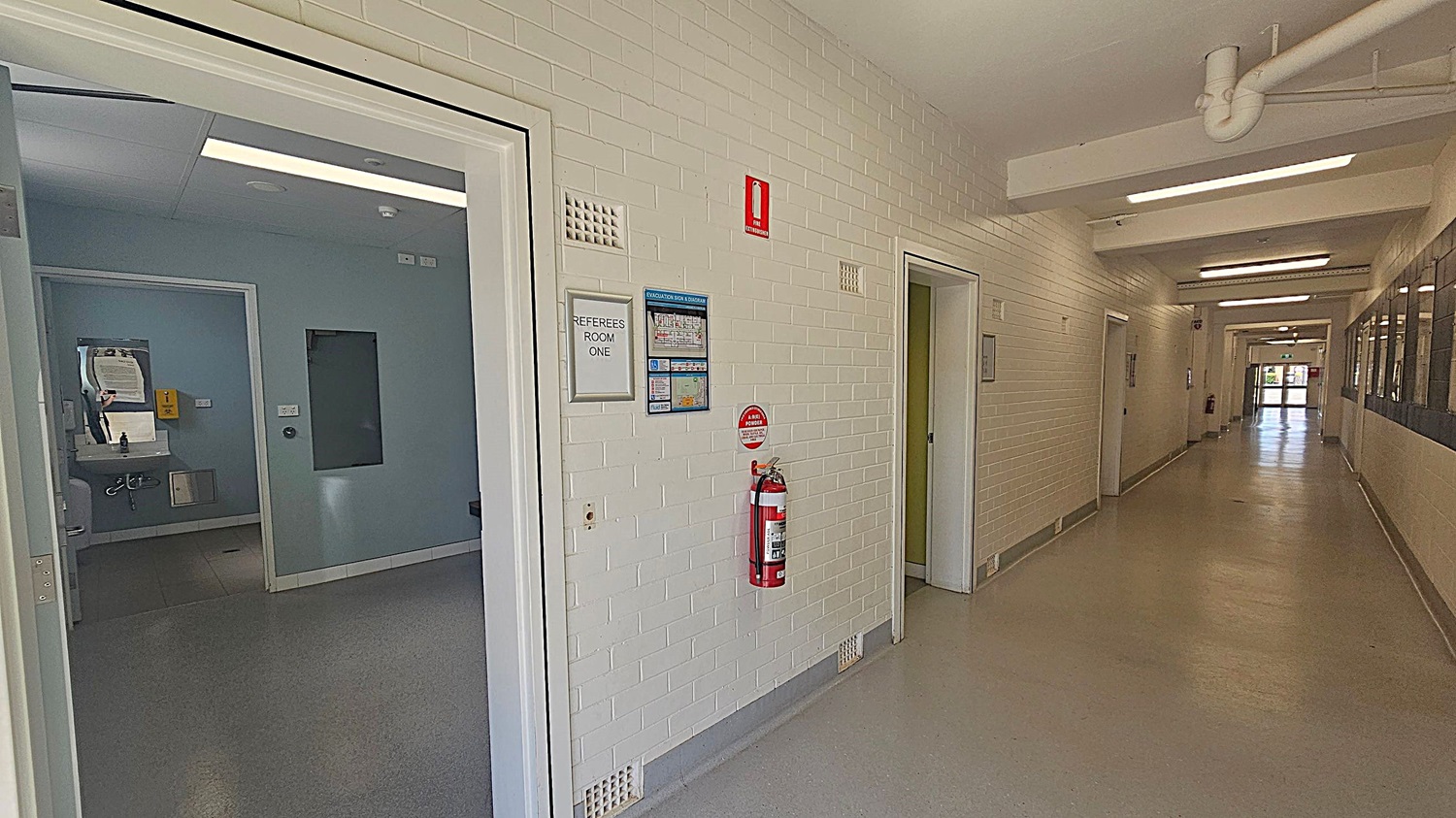 A corridor with white brick walls and a beige floor. On the left, an open door leads to "Referees Room One," with a visible sink and mirror inside. A fire extinguisher is mounted on the wall next to evacuation information. The corridor extends to the right, with several closed doors and windows along the walls. The ceiling has exposed pipes and fluorescent lights. At the end of the corridor, there is a glass door leading outside.