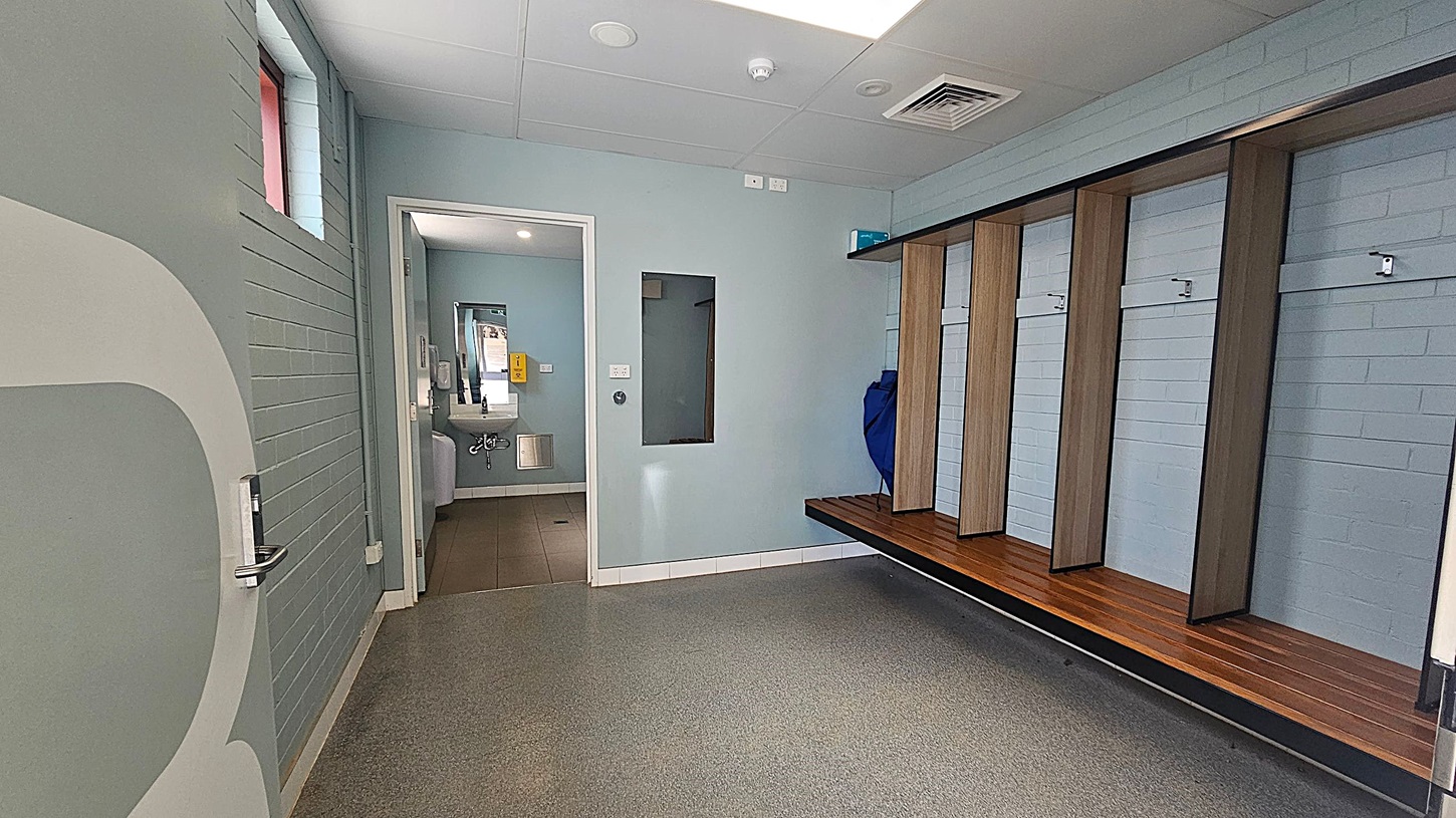 A changing room with a grey and green colour scheme. On the right, there are wooden benches with open cubbies above them. The floor is speckled grey and green. On the left, a partially open door leads to a corridor with a small window. In the centre, a doorway opens to a bathroom area with a visible sink and mirror. The ceiling is white with recessed lighting and ventilation panels.