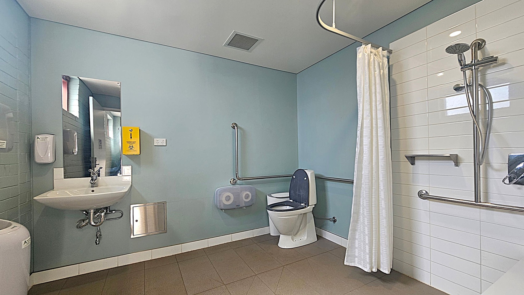 A spacious, accessible bathroom with light blue walls and a white ceiling. On the left, there's a sink with a mirror above it, a soap dispenser, and a yellow disposal box. The centre features a toilet with grab bars and a toilet paper dispenser. On the right, a shower area with a white curtain, handheld showerhead, and additional grab bars. The floor is tiled in a light brown colour.