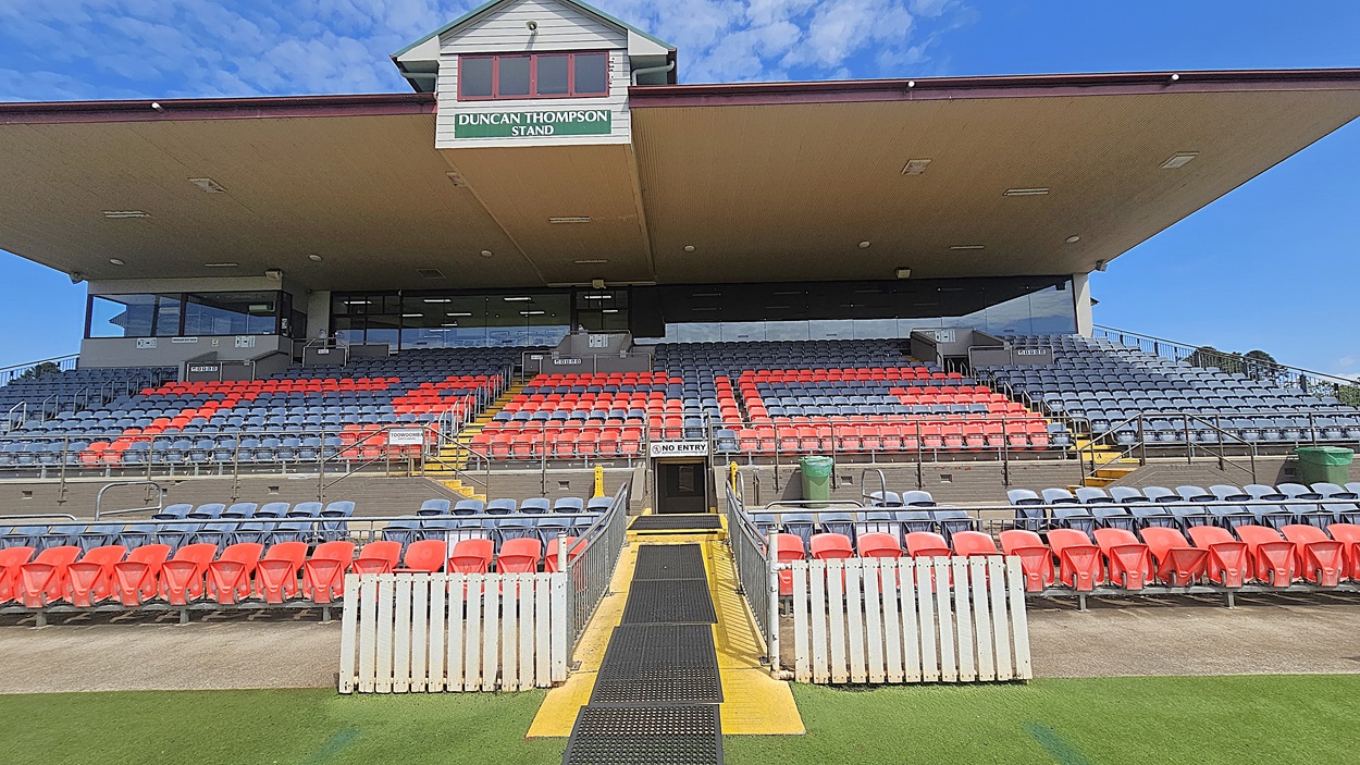 A sports stadium stand with a sign reading "Duncan Thompson Stand" at the top. The seating area features rows of red and grey seats. A central pathway with a "No Entry" sign leads up to the stand, flanked by white fencing. The stand has a large overhanging roof, and there are green bins on either side. The ground in front is covered with artificial turf.