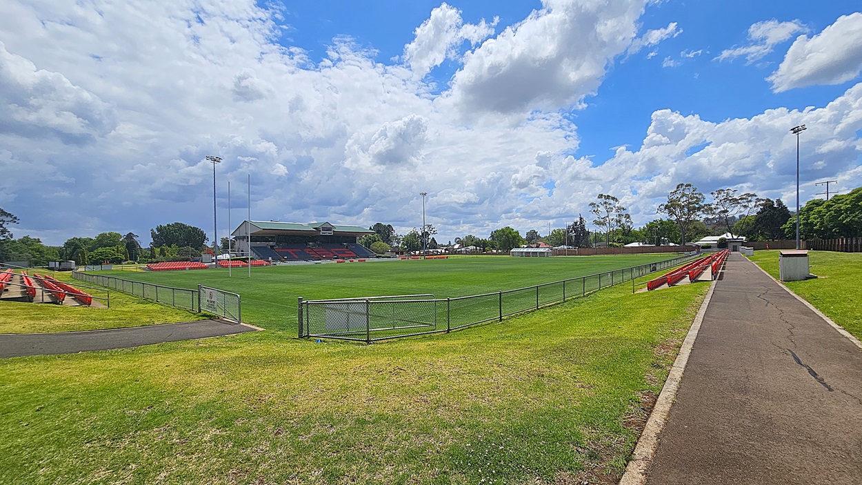 A sports field with a well-maintained green pitch is surrounded by a low metal fence. On the left, there are red seating stands, and a larger covered stand is visible in the background. On the right, a paved pathway runs alongside more red seating. Trees and a few buildings are in the distance, and tall floodlights are positioned around the field.