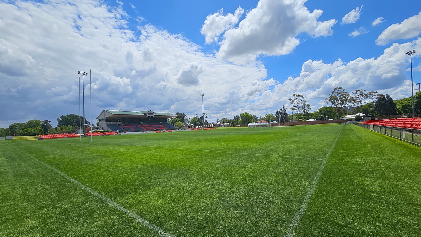A rugby field with green grass and white lines. On the left, there are rugby goalposts and a stand with red seats. In the background, a large building with a roof and more seating is visible. On the right, there are more red seats along the sideline, bordered by a fence. Trees and some small buildings are in the distance.