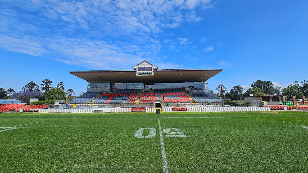 A sports stadium with a green field marked with a large "50" in the centre. The grandstand in the background has rows of red and blue seats, with a small building on top displaying a sign. Trees and additional structures are visible on either side of the grandstand. The sky is clear with some clouds.