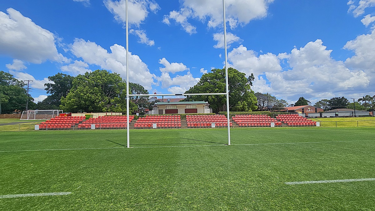 A rugby field with green grass and white goalposts in the foreground. In the background, there are rows of red seats forming a small grandstand. Behind the seats, there are trees and a building with a light-coloured facade. To the left, a football goal is visible. The sky is blue with scattered white clouds.
