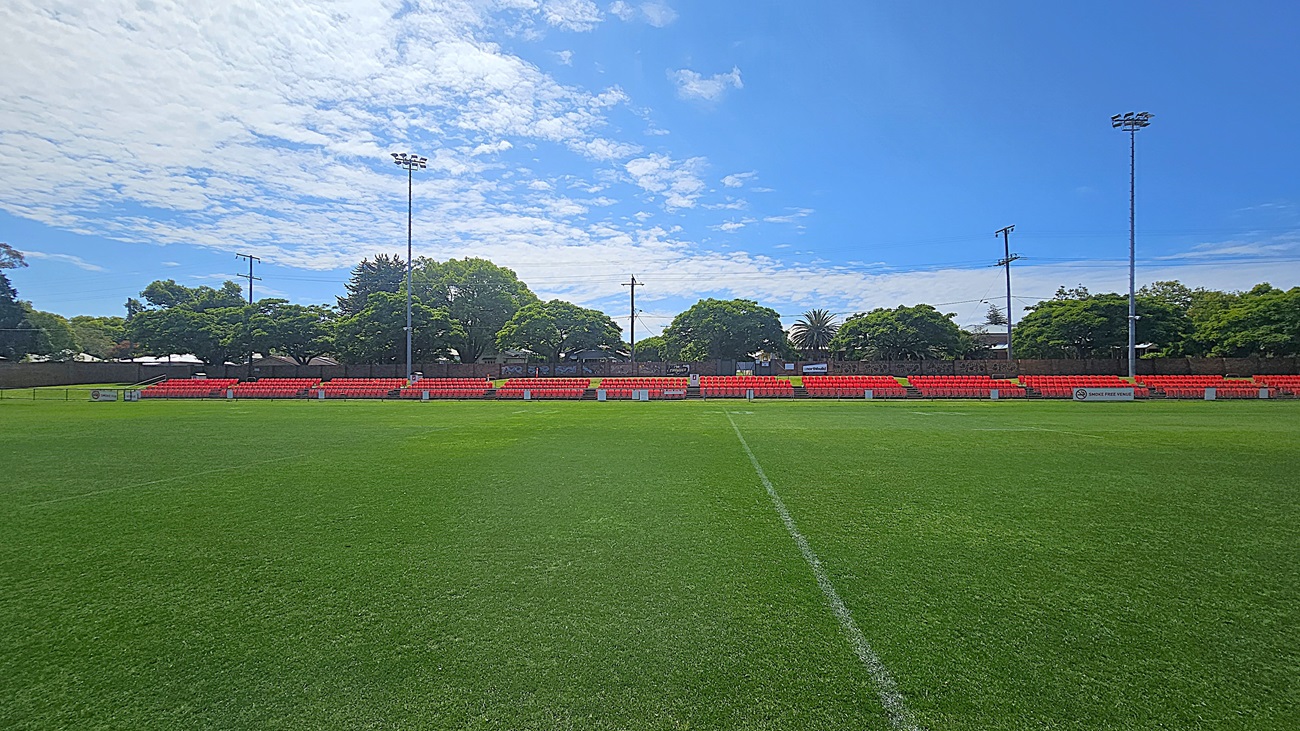 A football field with lush green grass. In the background, there are rows of red stadium seats, partially shaded by trees. Tall floodlights are positioned on either side of the seating area. The trees are dense and provide a natural backdrop, with some power lines visible above. The field is marked with white lines, indicating the playing area.