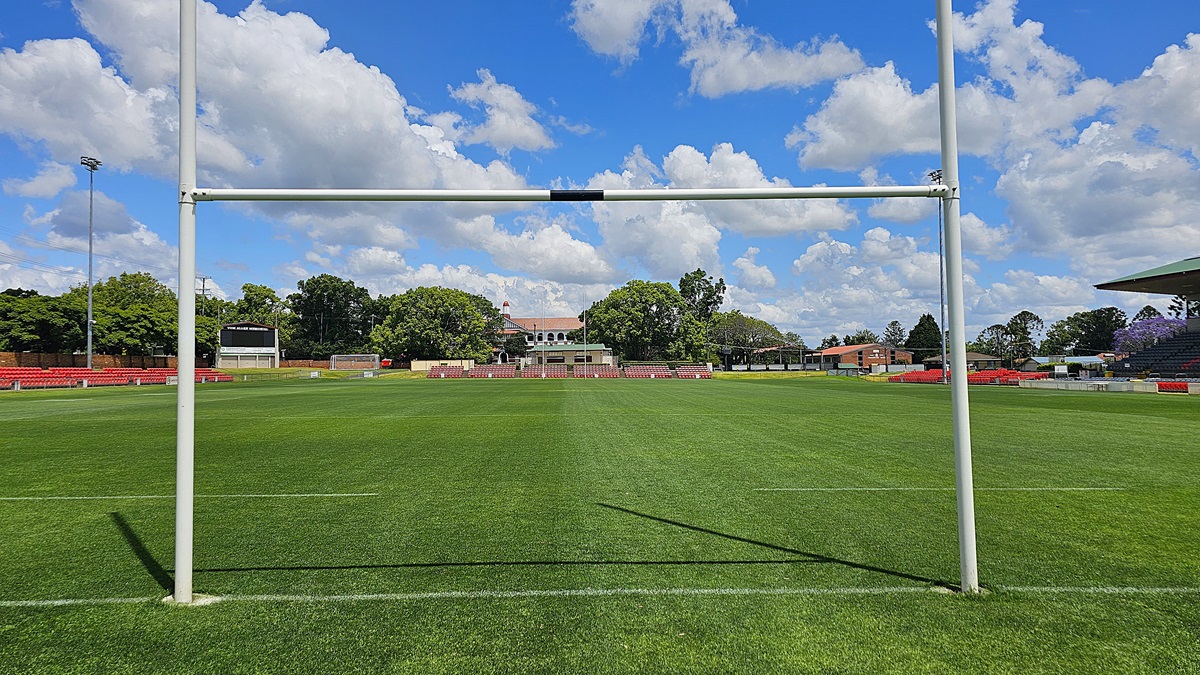 A rugby field with a goalpost in the foreground. The grass is neatly trimmed, and the field is surrounded by red seating. In the background, there are trees and a building with a red roof and arched windows. A scoreboard is visible to the left, and there are additional structures and trees on the right. The sky is filled with fluffy white clouds.