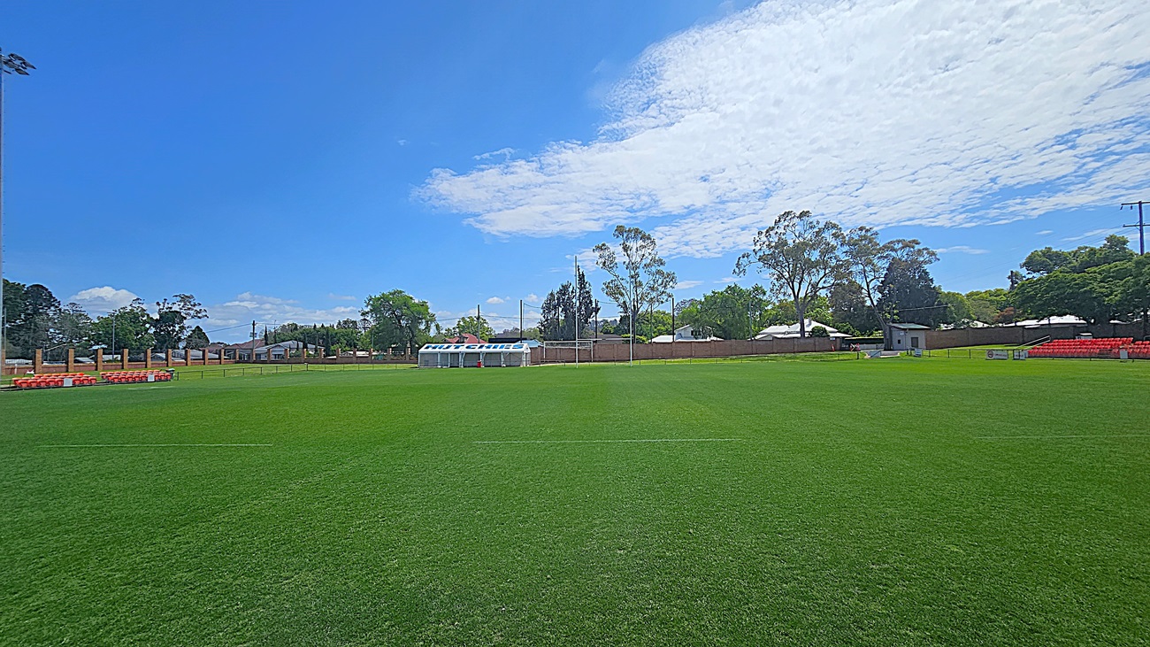 A green sports field with a row of orange seats on the left and right sides. In the centre background, there is a white shelter with a blue and white sign. Trees and a few buildings are visible in the background, along with a tall light pole on the left. The sky is mostly clear with some clouds.