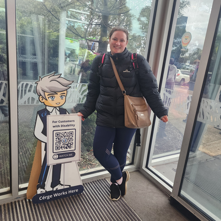 A woman in a black jacket stands beside a Cérge cartoon cutout with a QR sign.