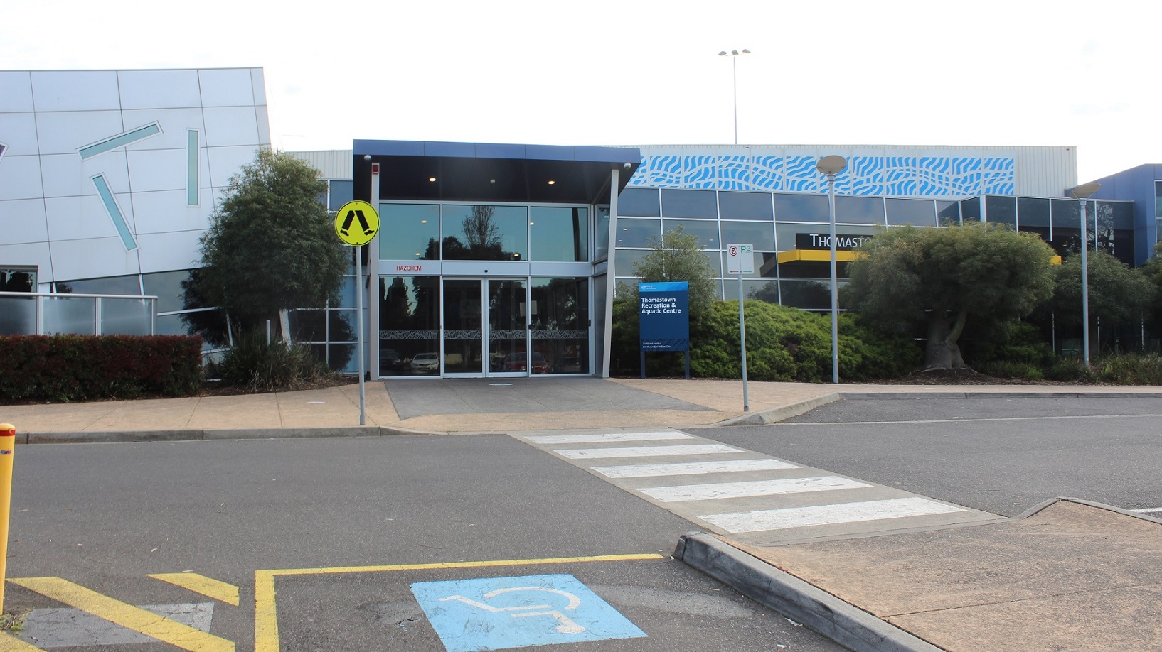 The Thomastown Recreation & Aquatic Centre building is a low-set glass and steel structure with blue and grey accents. On the right, there is a covered waiting area with a grey wooden chair close to a rubbish bin. In front of the facility, there are tall light poles and a sign on the façade that states "Thomastown Recreation Aquatic Center" in white on a black background with white trim. Grass and trees surround the building.
