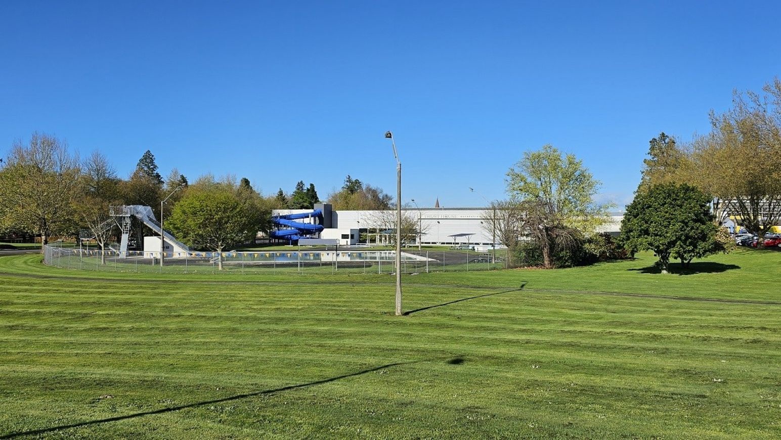 Distance view across park. Arrival and Parking. Trust House Recreation Centre – Masterton. A grassy field with neatly mowed lines stretches across the foreground. In the middle, a fenced area contains a swimming pool with a blue water slide. A grey building is visible behind the pool. Trees with green and brown foliage are scattered throughout the background. A single streetlamp stands in the centre of the image.