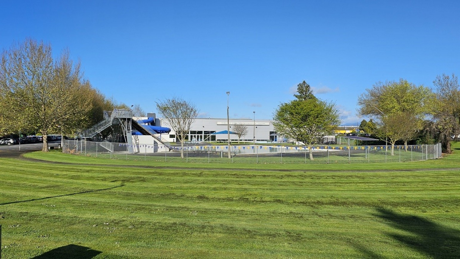 Across park field view. Arrival and Parking. Trust House Recreation Centre – Masterton. A grassy field with neatly mowed lines stretches across the foreground. In the middle, a fenced area contains a swimming pool with a blue water slide. A grey building is visible behind the pool. Trees with green and brown foliage are scattered throughout the background. A single streetlamp stands in the centre of the image.