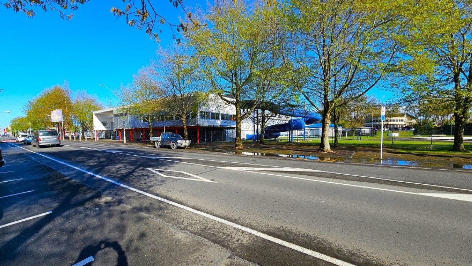 Street parking.  Arrival and Parking. Trust House Recreation Centre – Masterton.  A wide street with multiple lanes and a cycle lane on the left. Several cars are parked along the road. On the right, there is a modern building with large windows, partially obscured by a row of trees with green leaves. The pavement is wet, suggesting recent rain. The sky is clear and blue.