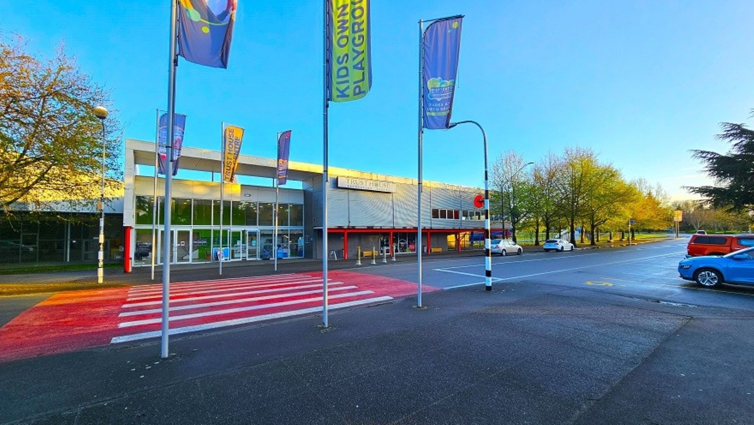 Pedestrian crossing and access. Arrival and Parking. Trust House Recreation Centre – Masterton. A modern building with a glass entrance is in the centre, featuring a red and white striped pedestrian crossing in front. Several tall flags with various designs are positioned along the crossing. To the right, a road with parked cars, including a blue and a red vehicle, extends into the distance. Trees with green foliage line the road, and the sky is clear.