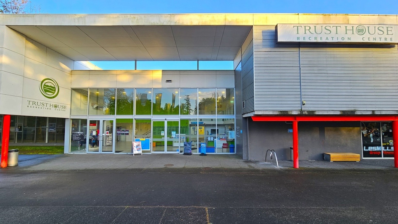 Entrance Façade. Arrival and Parking. Trust House Recreation Centre – Masterton.  A modern building with a large glass entrance in the centre, featuring automatic sliding doors. On the left, there's a logo and the name "Trust House" on the wall. The right side has a sign reading "Trust House Recreation Centre" above a section with horizontal metal cladding and red pillars. A small window displays "Les Mills" branding. The ground is paved, and there are bike racks and a bench near the entrance.