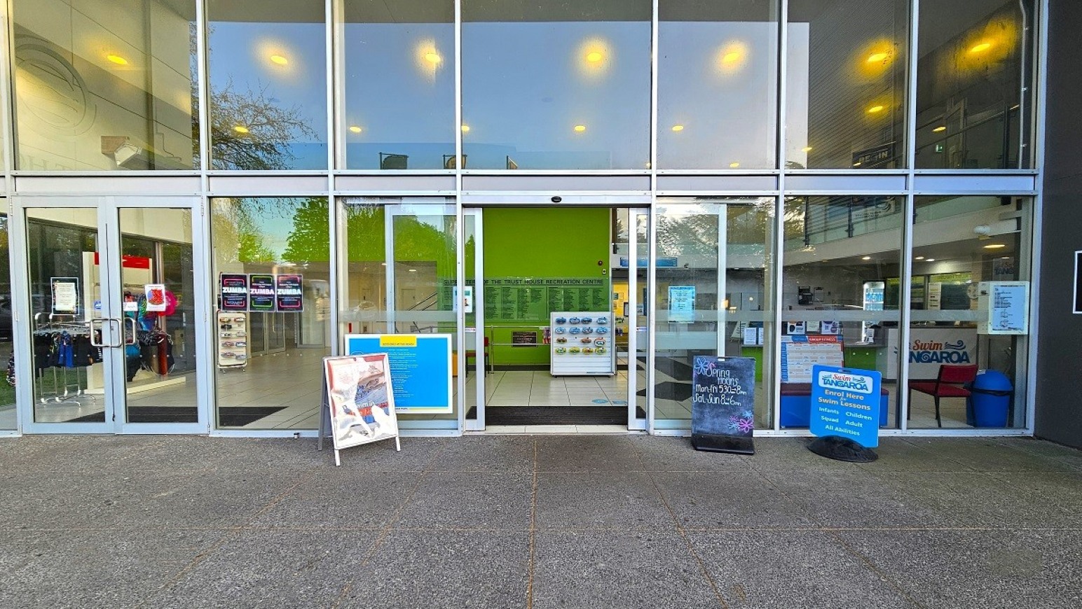 Automatic sliding door and step free entry.  Arrival and Parking. Trust House Recreation Centre – Masterton.  A modern building entrance with large glass doors and windows. On the left, there are posters and a clothing rack inside. In the centre, the doors are open, revealing a green wall and informational boards. On the right, there are more posters and a reception area with chairs. The floor is grey and the ceiling has bright lights.