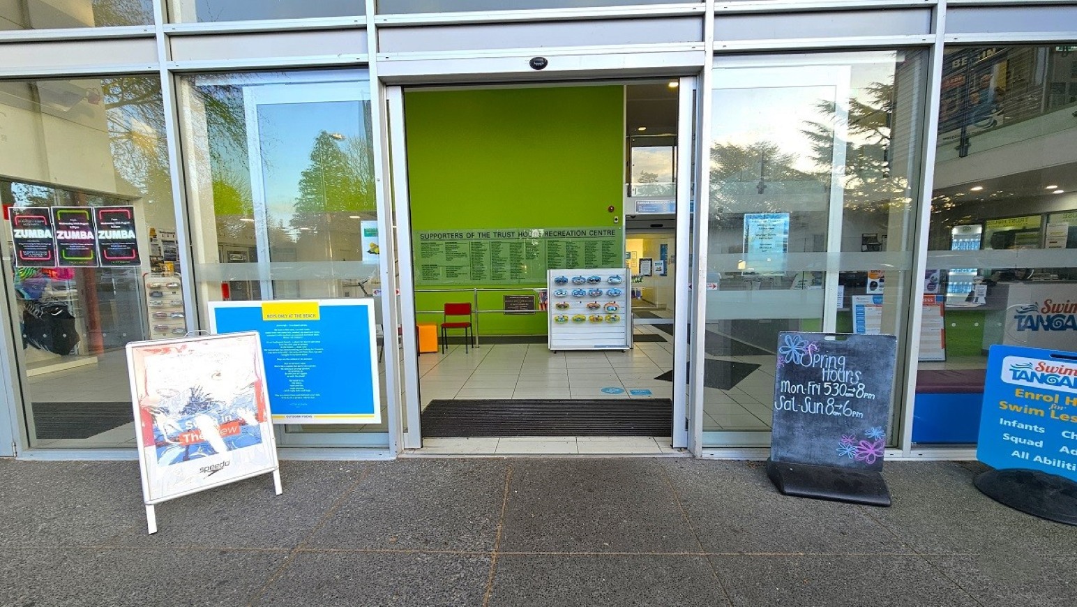 Automatic sliding door and step free entry.  Entrance and Reception.  Trust House Recreation Centre – Masterton.  A glass entrance with sliding doors leads into a building with a bright green interior wall. On the left, a white signboard with colourful graphics stands on the pavement. To the right, a blackboard displays opening hours, and another sign with blue and yellow text is visible. Inside, a small white display stand and a red chair are against the green wall.