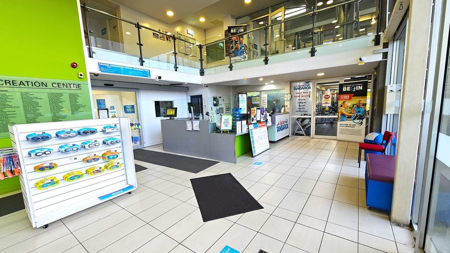 Reception and Retail.  Entrance and Reception.  Trust House Recreation Centre – Masterton.  A spacious indoor area with a tiled floor and a high ceiling. On the left, a green wall features a list of names and a display stand with swimming goggles. In the centre, a reception desk is surrounded by informational signs. To the right, glass doors lead to another room, with posters and a vending machine visible. Above, a glass-panelled balcony overlooks the space.