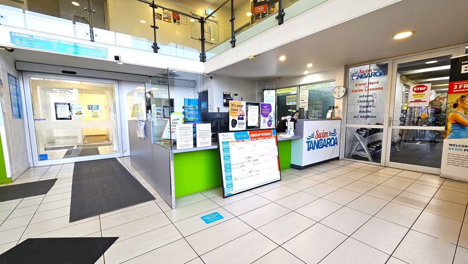 Reception, aquatic and gymnasium access.  Entrance and Reception.  Trust House Recreation Centre – Masterton.  A reception area with a green and white counter displaying various signs and information. To the right, a glass door with signage for swim lessons. Behind the counter, a wall clock and more informational posters. The floor is tiled in white, and there are dark mats leading to the entrance. Above, a glass railing indicates a second level. The ceiling is white with recessed lighting.