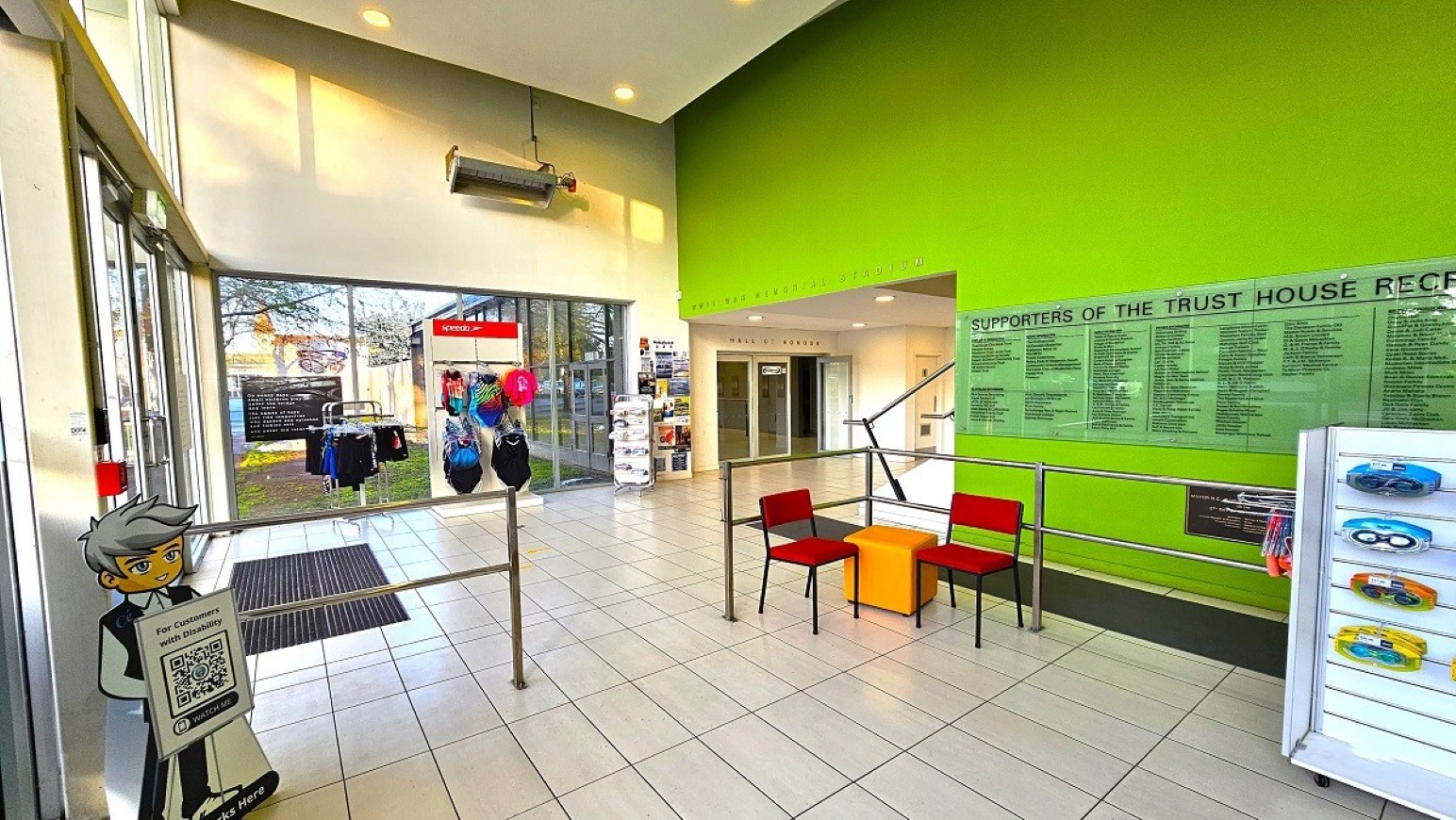 Reception foyer and stadium access.  Entrance and Reception.  Trust House Recreation Centre – Masterton.  Entrance area with large glass doors on the left, leading outside. A cartoon character cutout with a QR code stands near the door. In the centre, a tiled floor with a small seating area featuring two red chairs and a yellow cube. The right wall is bright green with a plaque listing supporters. A display case with swimming goggles is nearby.