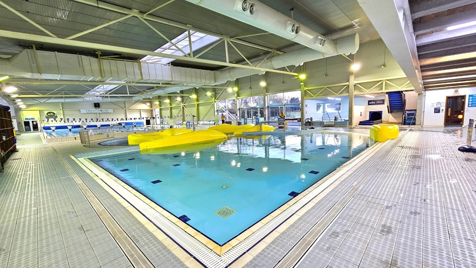 Pool deck on entry.  Aquatic Centre. Trust House Leisure Centre – Masterton. Indoor swimming pool area with a large pool in the centre featuring yellow inflatable obstacles. The pool is surrounded by a tiled deck. On the left, there is a lane pool with lane dividers. Large windows on the right allow natural light in. The ceiling is high with exposed beams and ductwork, and there are skylights. A staircase is visible in the background on the right, leading to an upper level.