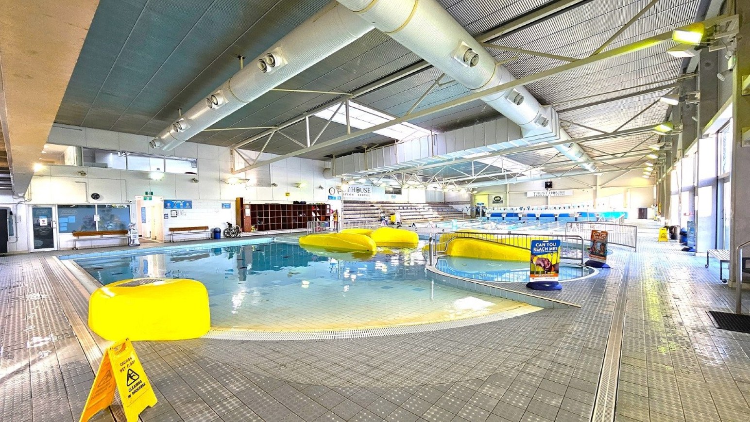 Pool deck step free entry.  Aquatic Centre. Trust House Leisure Centre – Masterton.  An indoor swimming pool area with a grey tiled floor and a high, metal-beamed ceiling. In the centre, there is a small pool with large yellow inflatable structures. To the right, a larger pool with lanes is visible. On the left, there are benches and a wall with windows. The right side features large windows allowing natural light in. A yellow caution sign is on the floor near the pool.