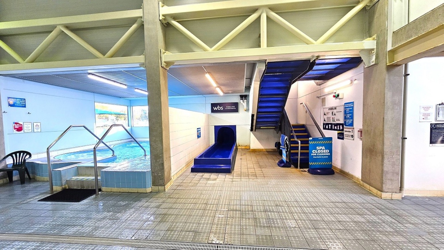 Spa Area.  Aquatic Centre. Trust House Leisure Centre – Masterton. On the left, there is an indoor swimming pool with metal handrails and a black chair nearby. The pool area is well-lit with overhead lights. In the centre, a blue slide leads into the pool. On the right, a blue staircase ascends, with a sign indicating the spa is closed. The floor is tiled, and the ceiling has exposed beams and lighting. Various signs and notices are displayed on the walls.