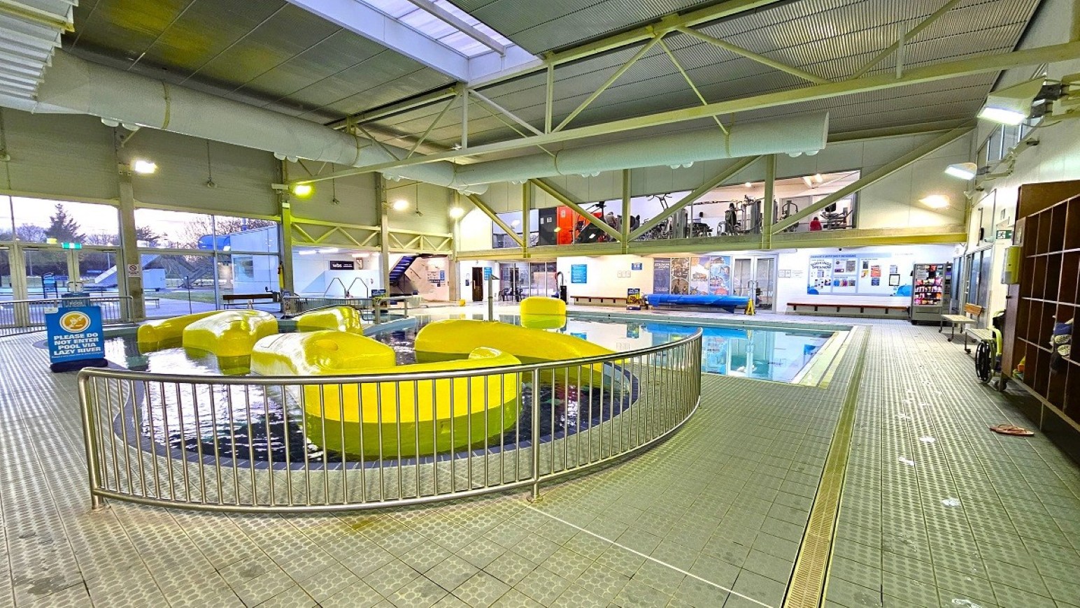 Lazy River. Aquatic Centre. Trust House Leisure Centre – Masterton. Indoor swimming pool area with a tiled floor and a high, metal-beamed ceiling. In the foreground, a circular pool with large, yellow floating structures is enclosed by a metal railing. To the right, a larger swimming pool is visible, with benches and a vending machine along the wall. The background features large windows and a mezzanine level with people visible.