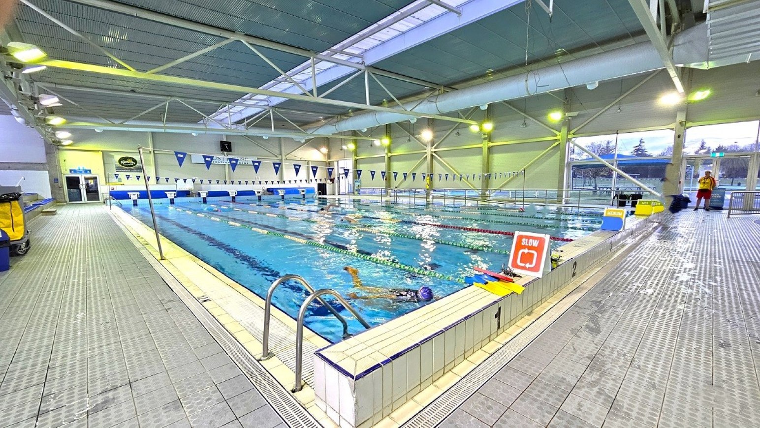 Pool deck 25m pool.  Aquatic Centre. Trust House Leisure Centre – Masterton. An indoor swimming pool with several lanes, each marked by floating lane dividers. Swimmers are in the water, and a "Slow" sign is visible at the pool's edge. The ceiling is high with exposed beams and lighting fixtures. On the left, there is a tiled walkway with a metal handrail leading into the pool. Large windows on the right allow natural light to enter, and a few people are standing near the windows.