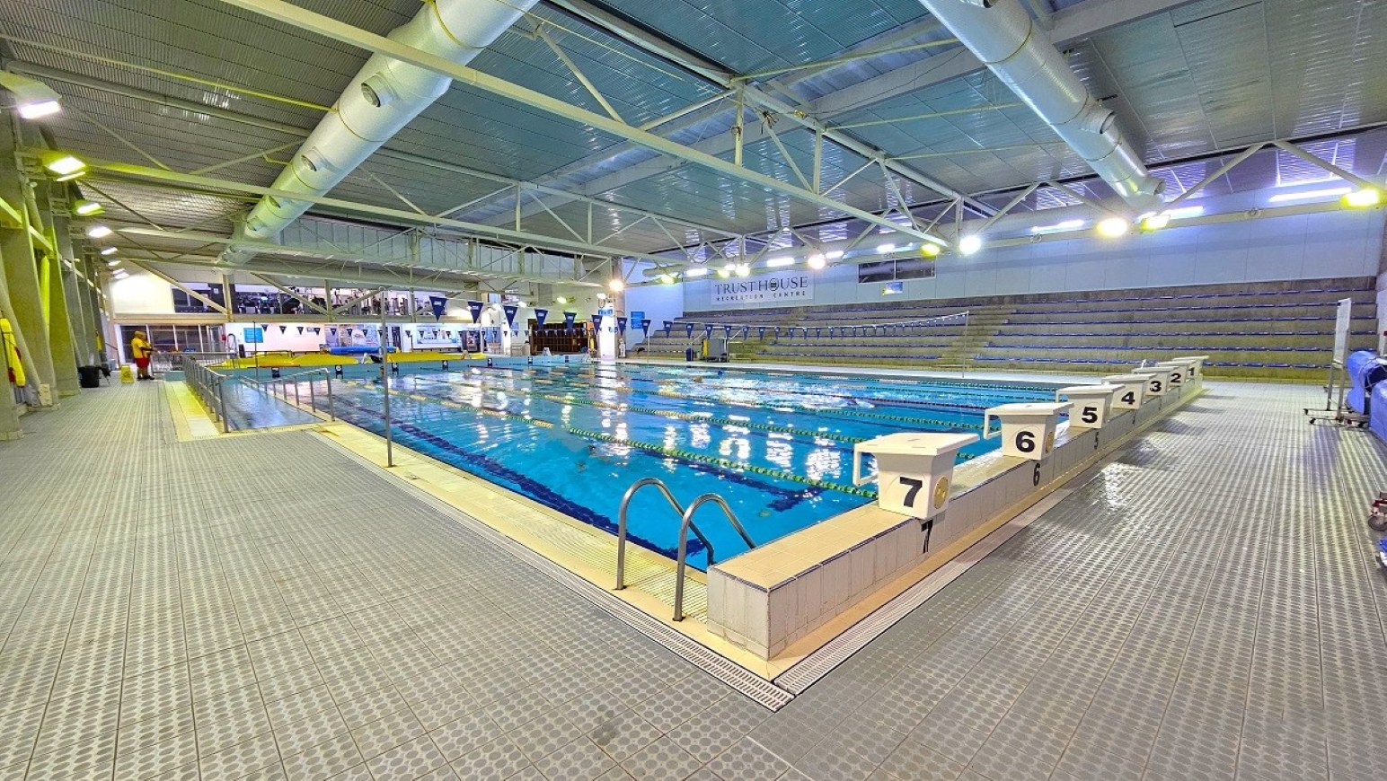 Reverse view 25m pool.  Aquatic Centre. Trust House Leisure Centre – Masterton. Indoor swimming pool with clear blue water, surrounded by a tiled deck. On the right, there are starting blocks numbered 2 to 7. The left side features a walkway with safety equipment on the wall. In the background, there are tiered seating areas for spectators. The ceiling is high with exposed beams and large ventilation ducts. Bright overhead lights illuminate the space.