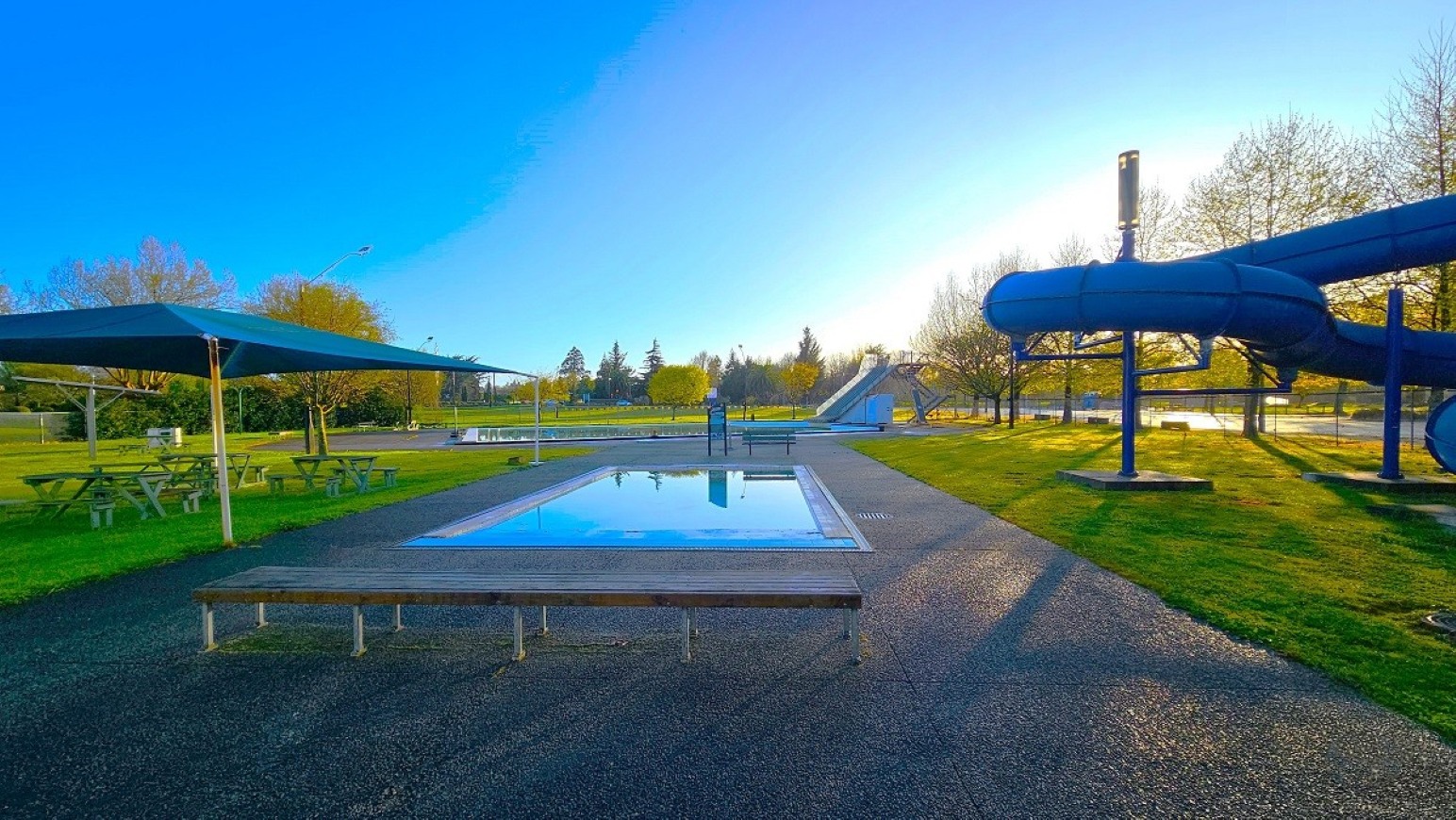 Outdoor pool.  Aquatic Centre. Trust House Leisure Centre – Masterton. A small rectangular pool is in the centre, surrounded by a paved area. On the left, there are picnic tables under a large shade structure. On the right, a blue water slide curves above the grass. In the background, trees and a larger slide are visible. The sky is clear and blue, and the grass is well-maintained.