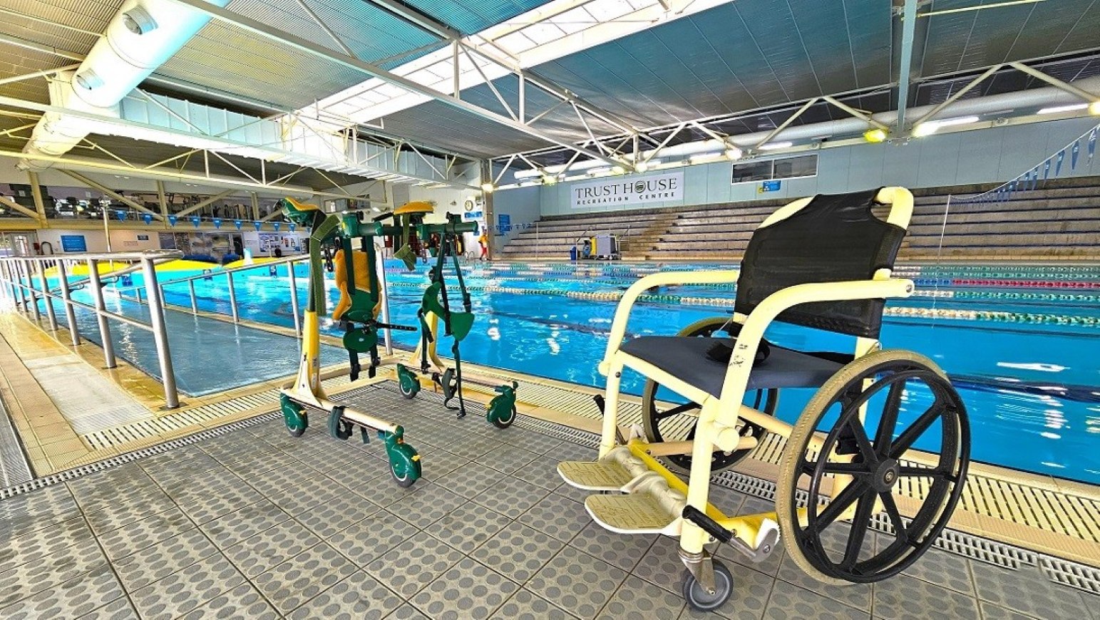 Aquatic wheelchair and ramp entry.  Aquatic Centre. Trust House Leisure Centre – Masterton. A swimming pool area with a wheelchair and mobility aids on the tiled floor in the foreground. The pool is in the centre, with clear blue water and lane dividers. On the right, there are tiered seating areas for spectators. The ceiling is high with exposed beams and lighting fixtures. The left side features a railing along the pool's edge.