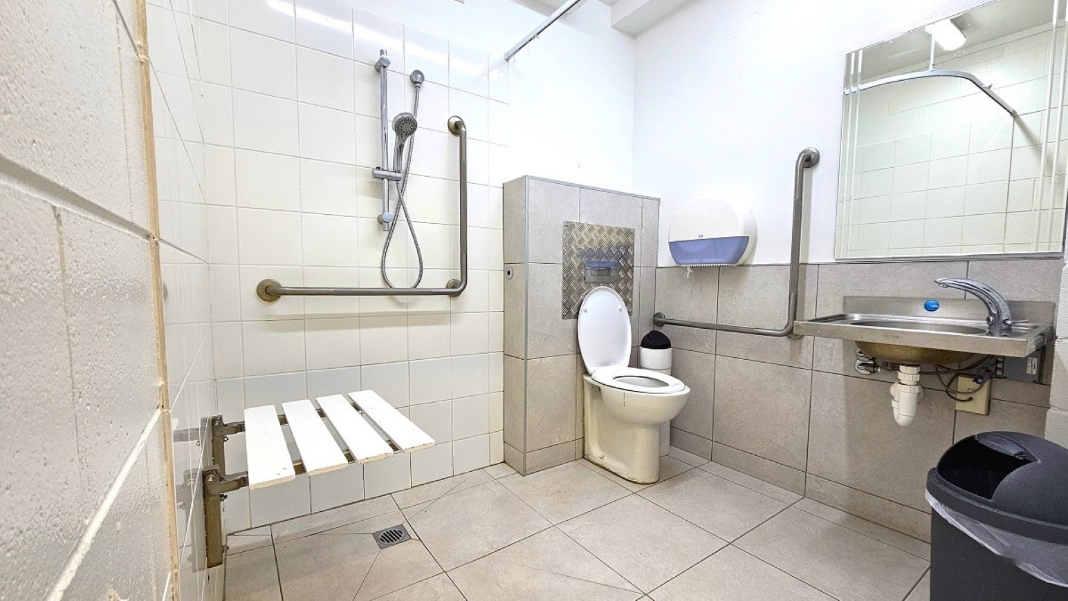 LH Accessible bathroom.  Aquatic Centre. Trust House Leisure Centre – Masterton. Accessible bathroom with white tiled walls and a light grey tiled floor. On the left, a shower area with a handheld showerhead and a foldable white seat. A horizontal grab bar is mounted on the wall. In the centre, a toilet with an open lid and a small bin beside it. On the right, a sink with a mirror above and a blue soap dispenser. Grab bars are installed near the toilet and sink.