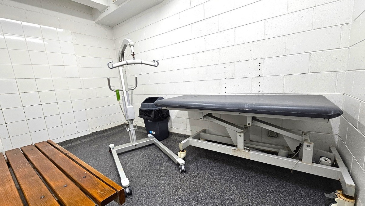 Change room and adult change table. Aquatic Centre. Trust House Leisure Centre – Masterton. A small room with white tiled walls and a grey speckled floor. On the right, there's a black cushioned examination table with a metal frame. In the centre, a white mobile hoist with a green handle is positioned next to a black bin. On the left, a wooden bench with a slatted design is visible. The ceiling is white with a slight overhang.