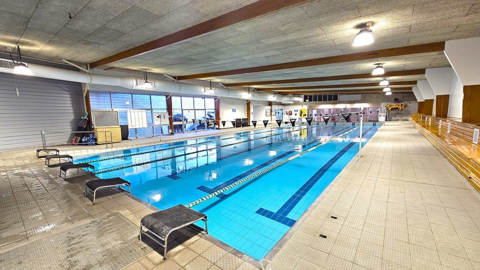 Pool deck. 25m Program Pool. Trust House Recreation Centre – Masterton. Indoor swimming pool with clear blue water, divided into lanes by floating ropes. On the left, there are several black diving blocks and a tiled floor area. The right side features a wide walkway with wooden benches and a railing. The ceiling is lined with exposed beams and hanging lights. Large windows on the left wall allow natural light to enter, and various equipment is stored near the windows.