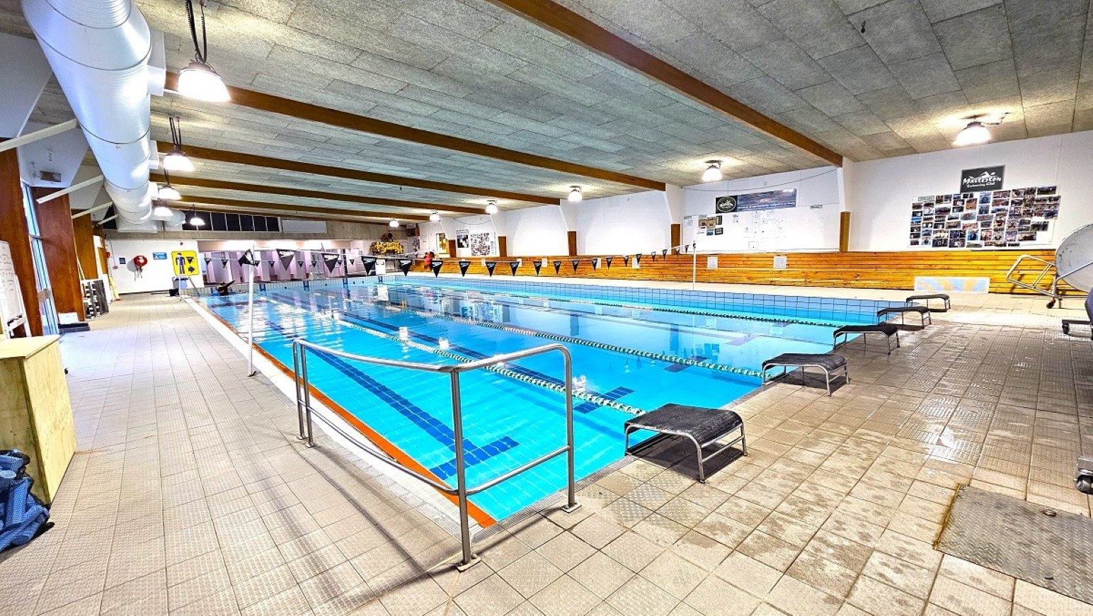 Reverse view pool deck.  25m Program Pool. Trust House Recreation Centre – Masterton. Indoor swimming pool with clear blue water, divided into lanes by floating ropes. On the left, a tiled walkway with metal railings and a large ventilation duct on the ceiling. The right side features wooden benches and walls adorned with posters and photographs. The ceiling is concrete with exposed beams and evenly spaced lights. The floor is tiled, and there are starting blocks at the pool's edge.