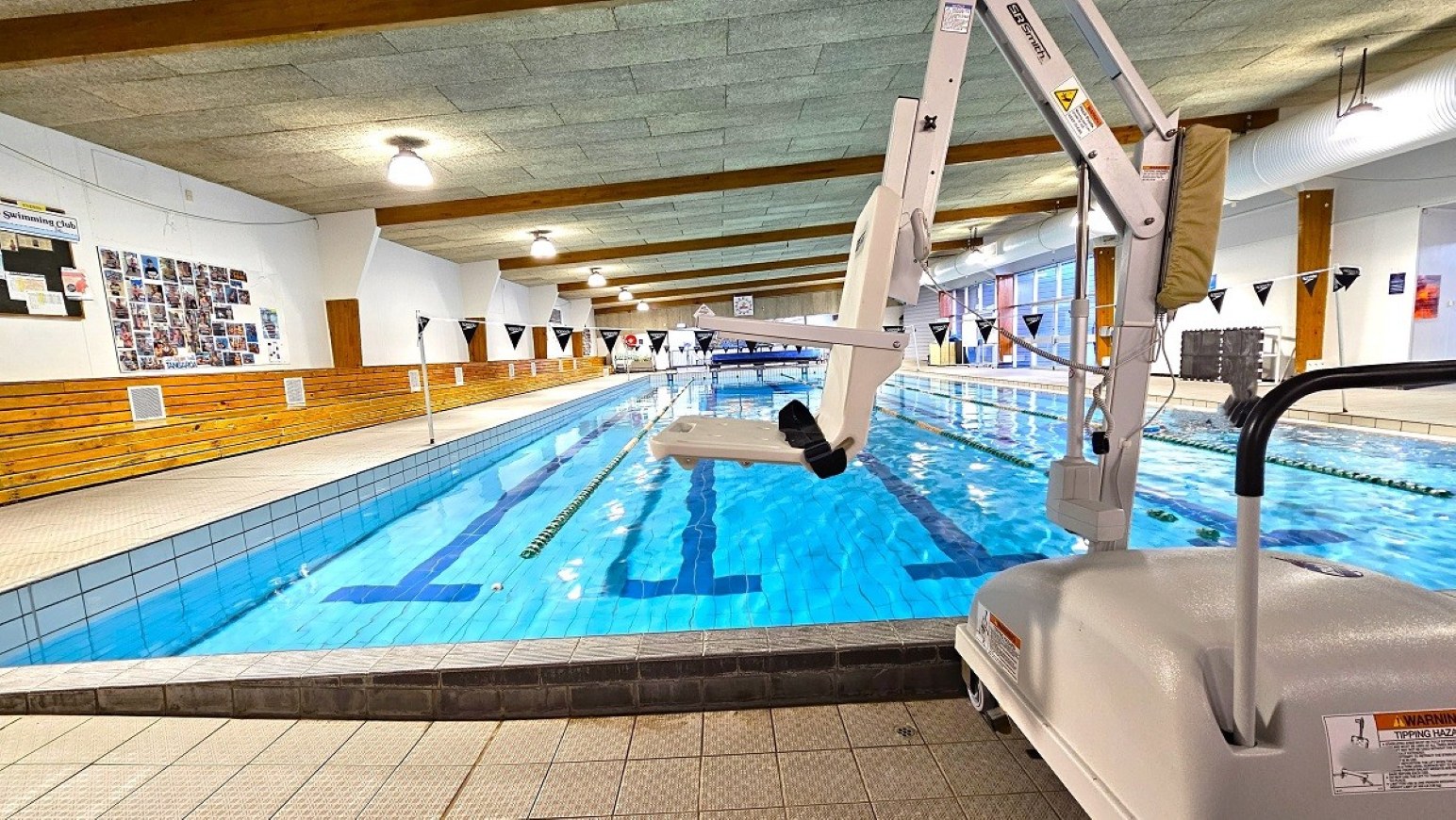 Pool hoist.  25m Program Pool. Trust House Recreation Centre – Masterton. An indoor swimming pool with clear blue water stretches into the distance. On the right, a pool lift is visible, designed for accessibility. The left side features wooden panelling and a wall with various photos and notices. The ceiling has exposed beams and hanging lights. The floor is tiled in a light colour, and triangular flags hang above the pool. Large windows on the right allow natural light to enter.