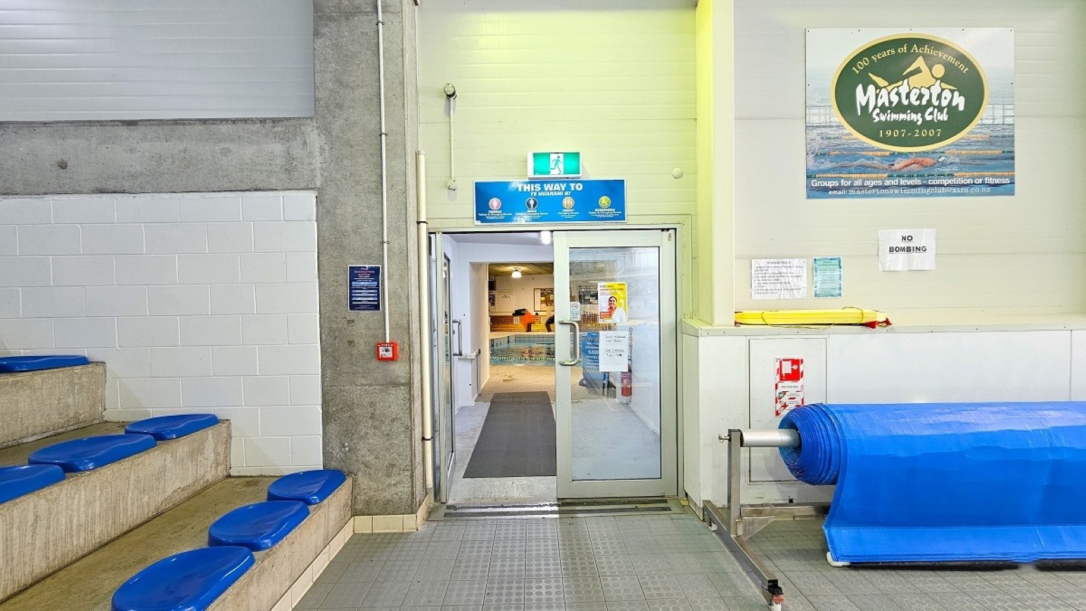 Program Pool Entrance Door. 25m Program Pool. Trust House Recreation Centre – Masterton.  A swimming pool area with tiered seating on the left, featuring blue cushioned seats. In the centre, there are glass double doors with a sign above indicating directions. On the right, a wall displays a poster for the Masterton Swimming Club and a "No Bombing" sign. Below, a blue pool cover is rolled up. The floor is tiled, and the ceiling is not visible.