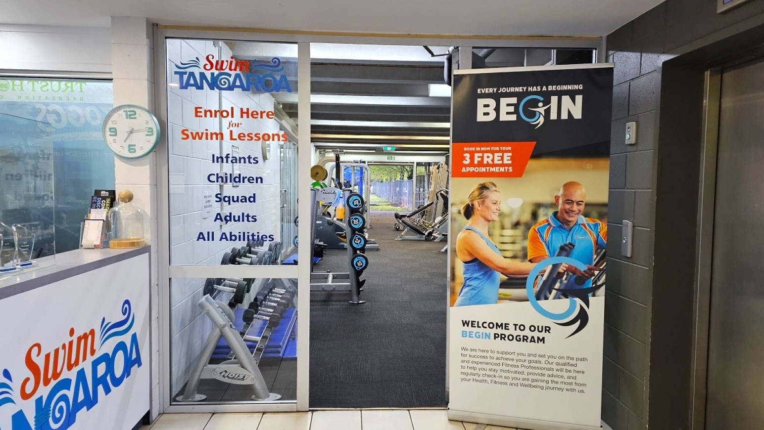 Ground level gym entry doorway.  Gymnasium Ground Level.  Trust House Recreation Centre – Masterton.  On the left, there's a reception counter with the logo "Swim Tangaroa" and a clock above it. A glass door displays information about swim lessons for various age groups and abilities. In the middle, a gym area with exercise equipment is visible through the glass. On the right, a large banner promotes a fitness programme with an image of two people exercising.