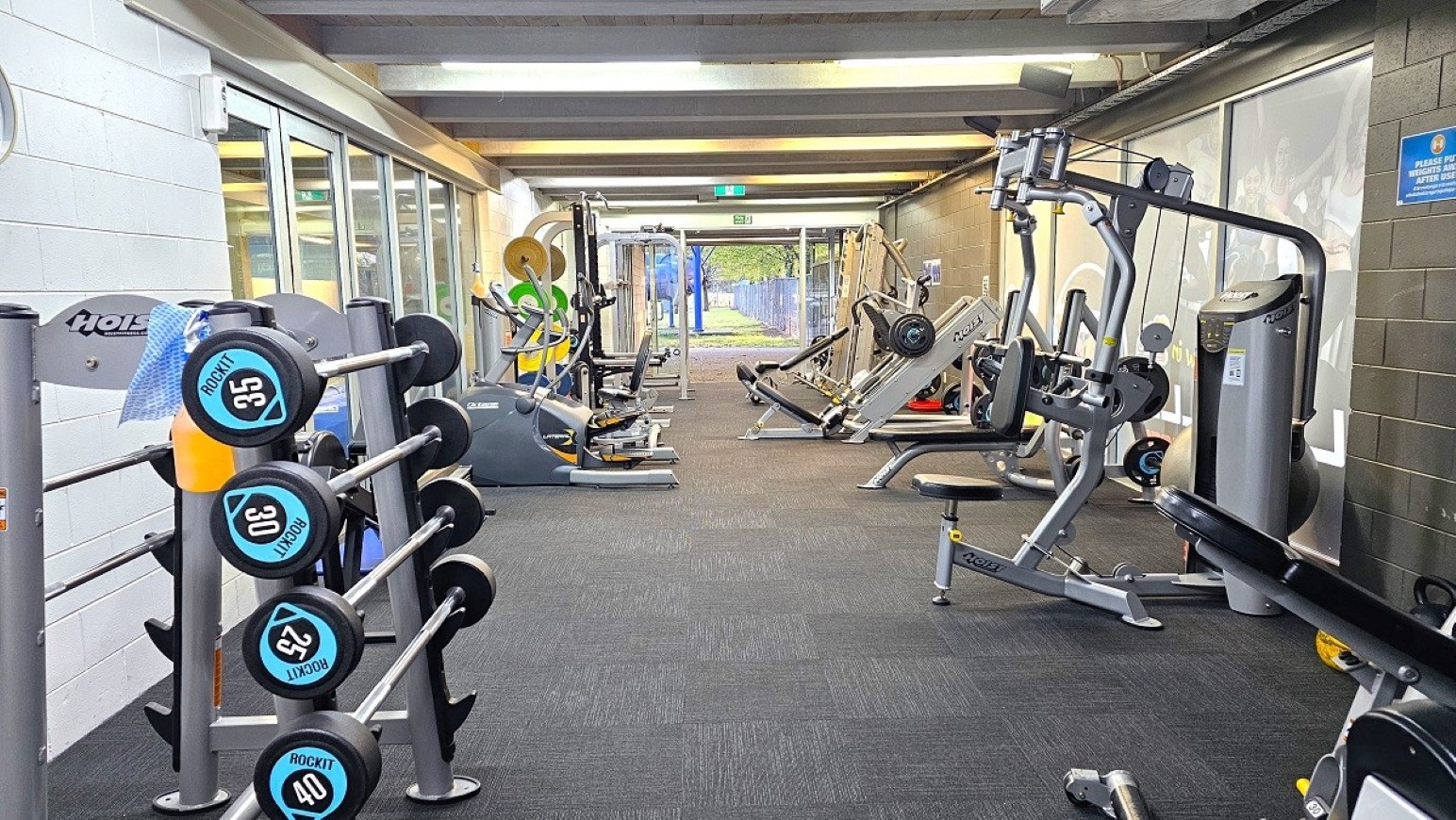 Resistance weights.  Gymnasium Ground Level.  Trust House Recreation Centre – Masterton. A gym with various exercise equipment. On the left, there is a rack of dumbbells with weights ranging from 30 to 40. The right side features a range of machines, including a leg press and a multi-exercise machine. The floor is carpeted in dark grey, and the ceiling has exposed beams with fluorescent lighting. The background shows a glass door leading outside, with a view of greenery.