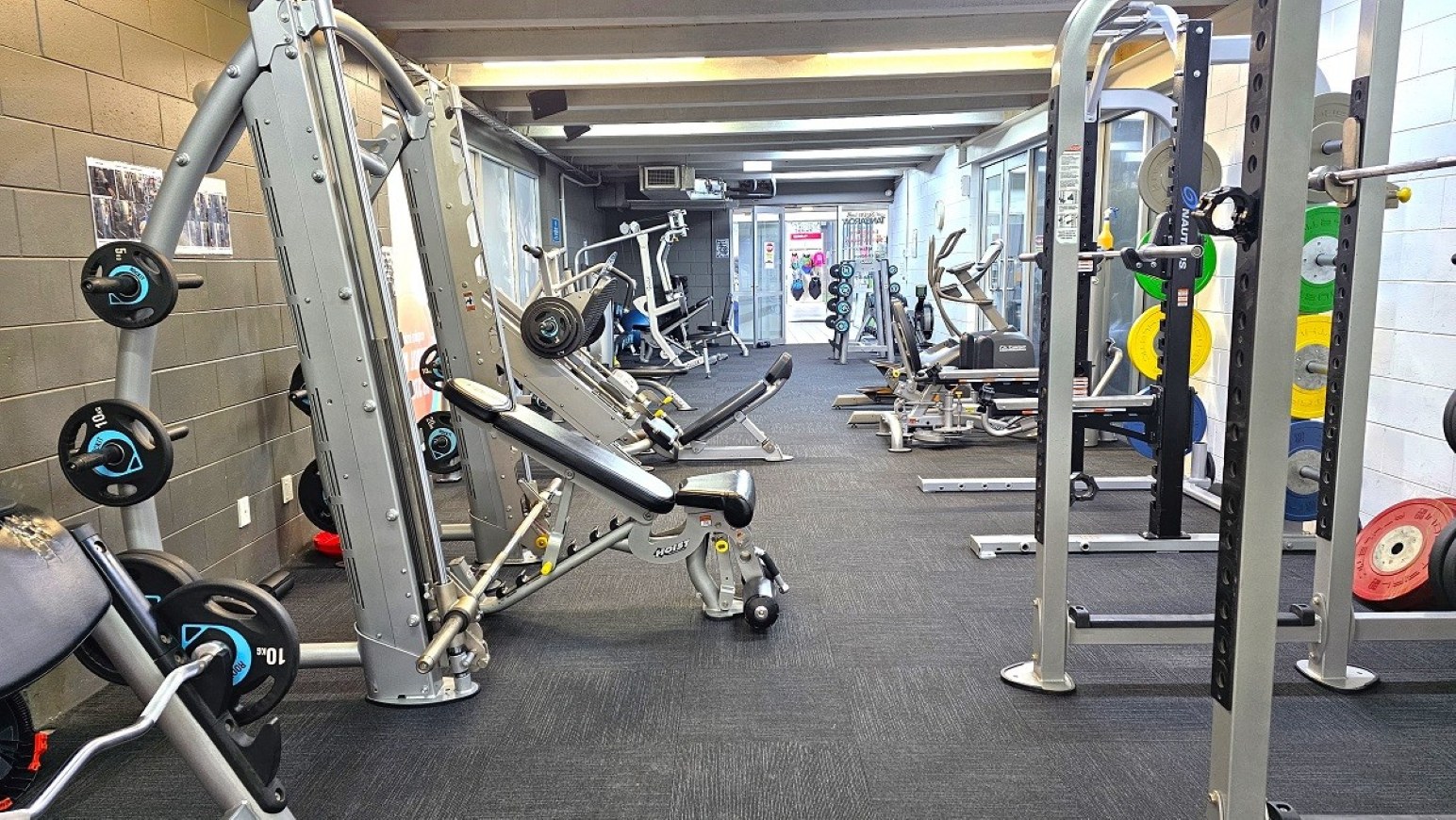Reverse view resistance weights.  Gymnasium Ground Level.  Trust House Recreation Centre – Masterton. A gym with various exercise equipment. On the left, there are weight machines and benches with weights. The floor is carpeted in dark grey. On the right, there are more weight racks with colourful weight plates. In the background, there are additional machines and a glass door leading to another area. The ceiling has exposed beams and lighting fixtures.